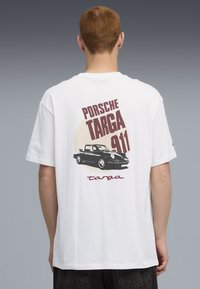 Hvid bomulds T-shirt med et sort Porsche 911 Targa grafik på ryggen, med vinrødt tekst og lys cremefarvet cirkulært baggrund.