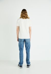 Camiseta branca de gola redonda, jeans de ganga azul e ténis pretos. Tecido suave, corte descontraído, design básico com vista traseira exibida.