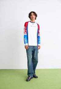 Langærmet t-shirt i hvid med røde og blå flammer, blå ærmer og logo. Kombineret med løstsiddende blå jeans og sorte sneakers.