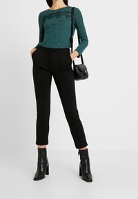 Turkusowa bluzka z długim rękawem z koronkowymi detalami przy dekolcie, w połączeniu z czarnymi eleganckimi spodniami i czarnymi botkami na obcasie.
