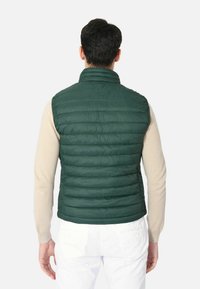 Gilet verde imbottito con colletto alto, caratterizzato da una texture trapuntata. Indossato sopra una camicia a maniche lunghe beige, in tessuto leggero e design affinato.