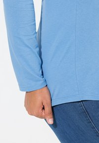 Světle modrá košile s dlouhými rukávy a kulatým výstřihem, doplněná tmavě modrými džíny. Tkanina vypadá hladce a elasticky.