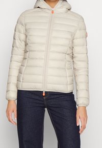 Helle beige Pufferjacke mit Reißverschluss vorne, Kapuze und elastischen Bündchen. Mit horizontalem Steppmuster und orangefarbener Reißverschluss-Zuglasche.