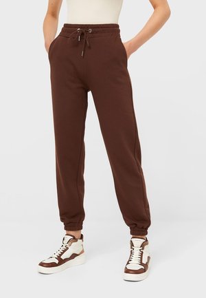 Persona vistiendo pantalones jogger marrones con cordón y puños elásticos, y zapatillas altas blancas y marrones, de pie con las manos en los bolsillos.