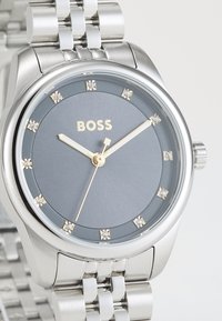 Montre en acier inoxydable avec un cadran gris, des accents dorés et des index ressemblant à des diamants. Elle dispose d'un bracelet en métal à maillons et d'un design de boîtier rond.