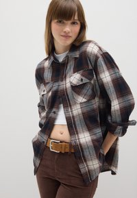 Gepofte flanellen shirt in marineblauw, bruin en crème met twee borstzakken en opgerolde mouwen. Gerefereerd over een witte top en gecombineerd met een paar bruine broek en riem.