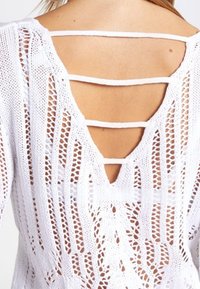 Haut en maille blanche présentant un motif en maille ouverte détaillé, un dos en V et plusieurs sangles horizontales à l\'arrière supérieur.