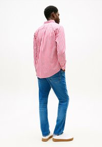 Camicia a maniche lunghe a strisce rosse e bianche con colletto a bottoni, abbinata a jeans blu e scarpe da ginnastica bianche. Dettagli di cucitura visibili.