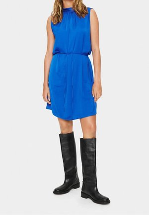 Heldere blauwe mouwloze jurk met een geplooide halslijn en een verstelbare taille, gecombineerd met knielange zwarte leren laarzen.