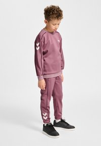 Paarse sportieve set bestaande uit een sweatshirt met ronde hals en witte chevrons op de mouwen, aangevuld met bijpassende afgezakte broeken. Gestyleerd met zwarte sneakers.