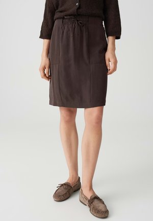 Donna che indossa una gonna marrone scuro fino al ginocchio con tasche, un maglione lavorato a maglia scuro e scarpe stile mocassino color taupe, in piedi su uno sfondo chiaro.
