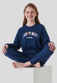 Blue cotton long-sleeve pajama shirt with "HAPPY MIND everyday" text, paired with matching blue pants, model sitting cross-legged on a white pedestal.