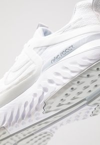 Białe buty Nike React mają siateczkową cholewkę, grubą teksturowaną podeszwę z wyraźnymi wzorami oraz markowe akcenty w jasnoszarym kolorze.