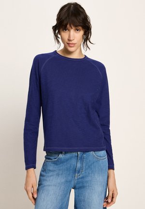 Junge Person mit dunklem Haar, die ein langärmeliges, navyblaues Shirt und hellblaue Jeans trägt, steht vor einem schlichten, hellen Hintergrund.