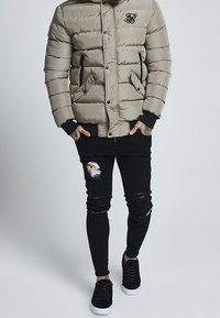 Beige padded jacket with a logo, black ripped jeans, and black sneakers. Jacket features snap buttons, pockets, and a textured finish.