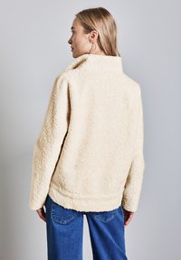 Veste en fausse fourrure crème avec un col haut, coupe décontractée et surface texturée. Associée à un jean bleu, vue de dos.