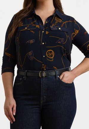 Kvinde iført en navy slå-om skjorte med guldkæde- og våbenmønstre, mørke jeans og et sort bælte med spænde i guld, hånd i lommen.