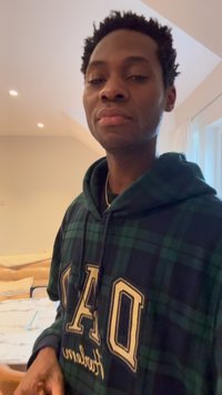 Man wearing green and black checkered hoodie with large "OPA" letters, standing indoors near a table with a glass and straw on it.