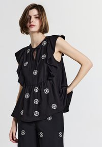 Donna con capelli corti castani che indossa un top senza maniche nero con ricami floreali bianchi e spalline arricciate, abbinato a pantaloni neri coordinati.