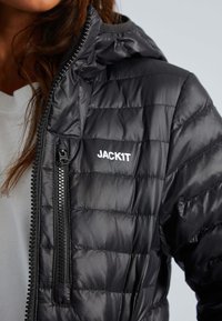 Zwarte pufferjack met klein wit logo "JACKIT," uitgerust met een ritssluiting en ribbelrand. De textuur is glad en licht glanzend.