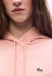 Ženska, ki nosi roza kapuco z majhnim zelenim logotipom krokodila na prsih, prikazuje ustnice in del dolgih rjavih las.