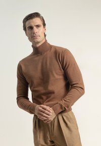 Hombre en suéter de tortuga marrón y pantalones beige de pie con las manos entrelazadas, mirando ligeramente hacia un lado contra un fondo simple.