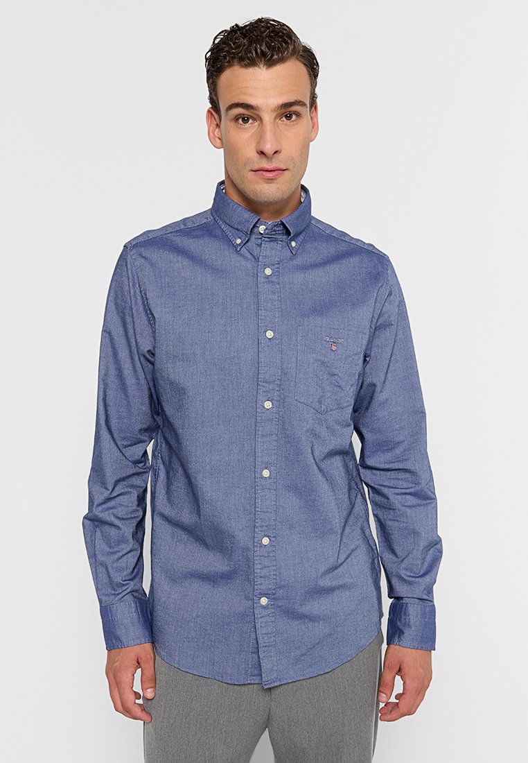 Hombre con una camisa azul de botones y un bolsillo en el pecho, de pie contra un fondo claro y liso, mirando hacia adelante.