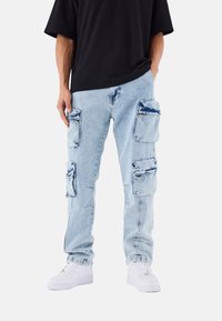 Pantalones cargo de mezclilla azul claro con múltiples bolsillos con cremallera, corte relajado y dobladillos enrollados. Combinados con zapatillas blancas y una camisa negra.