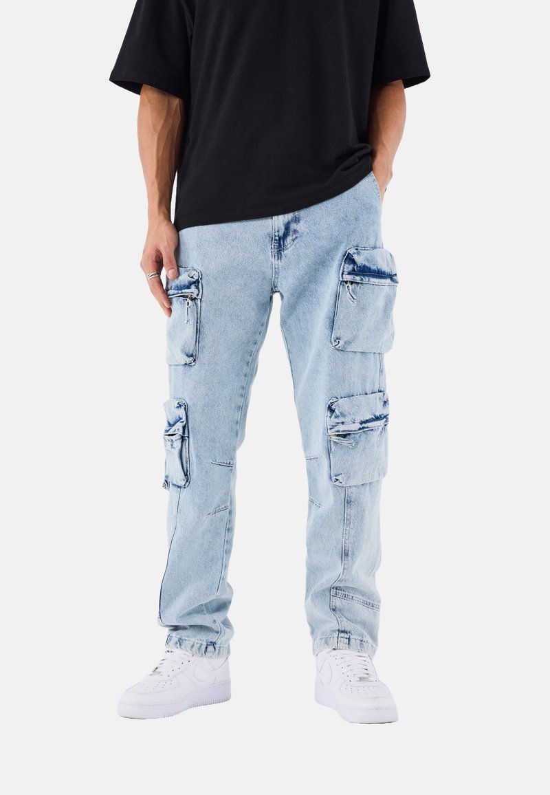 Pantalones cargo de mezclilla azul claro con múltiples bolsillos con cremallera, corte relajado y dobladillos enrollados. Combinados con zapatillas blancas y una camisa negra.