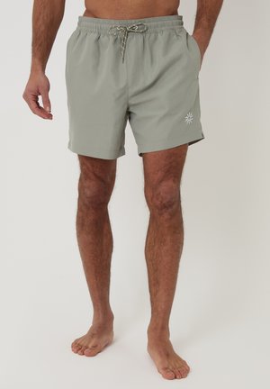 Mand iført lysegrå badebukser med snøre i taljen og sol-emblem på venstre ben, stående barfodet mod en ensfarvet baggrund.