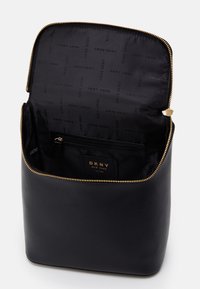 Czarna torba DKNY o teksturowanej powierzchni i złotym zamku błyskawicznym. Wnętrze wyściełane materiałem z logo DKNY oraz kieszenią na zamek. Kwadratowy kształt.