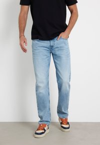 Jeans dritti blu chiaro con sbiaditure e leggeri segni di usura, abbinati a sneaker arancione e blu navy. T-shirt nera indossata sopra.