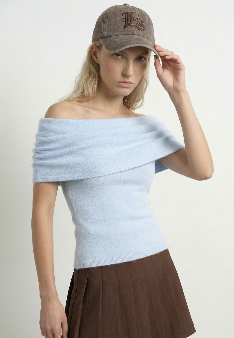 Jeune femme portant un haut bleu clair à épaules dénudées, une jupe plissée marron et une casquette marron, touchant la visière de la casquette avec une expression sérieuse.