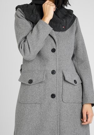 Kobieta ubrana w szary płaszcz zapinany na guziki z dużymi klapowymi kieszeniami oraz czarny dziergany szalik okrywający szyję i podbródek.