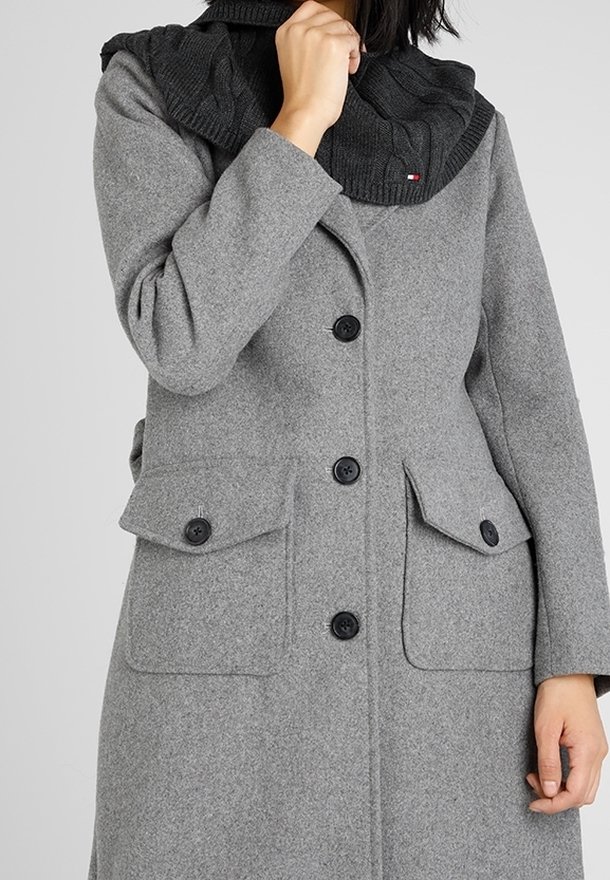 Femme portant un manteau gris boutonné avec de grandes poches à rabat et une écharpe noire en maille couvrant son cou et son menton.