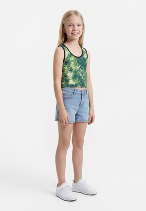 Fiatal lány áll, zöld levelekkel díszített ujjatlan felsőt, világos farmerrövidnadrágot és fehér tornacipőt visel, mosolyog egy sima fehér háttér előtt.