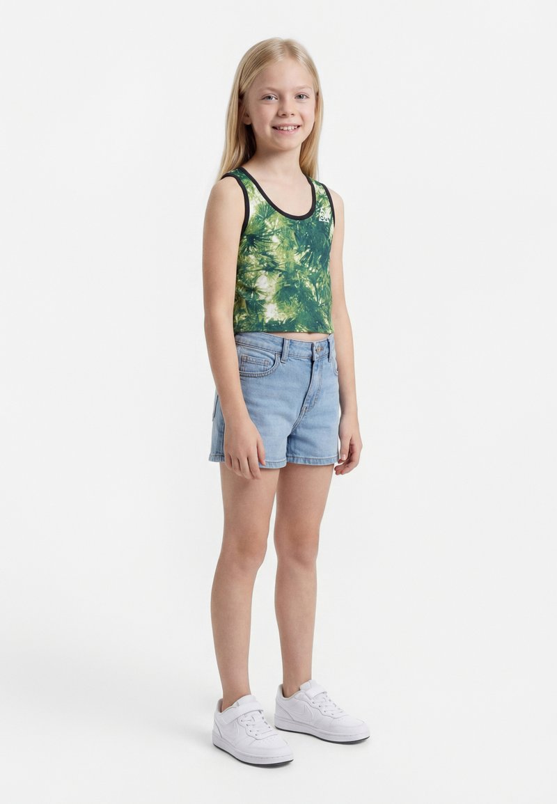 Noor tüdruk seisab, seljas roheline lehemustriga topp, heledad teksapüksid ja valged tossud, naeratab ühevärvilise valge tausta ees.