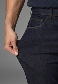 Dunkelblaue Denim-Jeans mit klassischem Fünf-Taschen-Design. Weiche Textur, gerader Schnitt, sichtbare Nähte und eine Hand, die den Stoff in der Nähe der Tasche greift.
