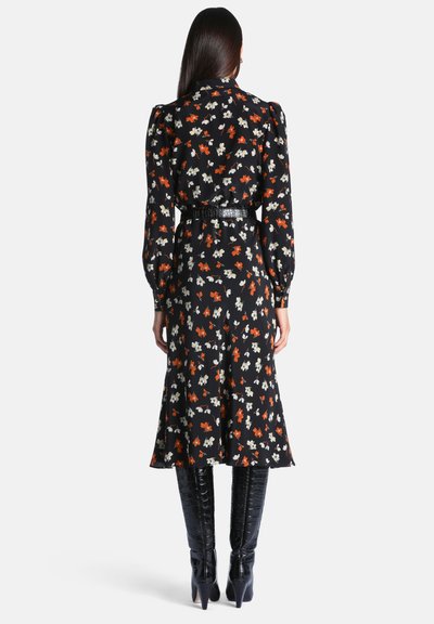 Femme debout, vue de dos, portant une robe midi noire à fleurs avec des manches longues et des bottes noires à talons montant jusqu'aux genoux.