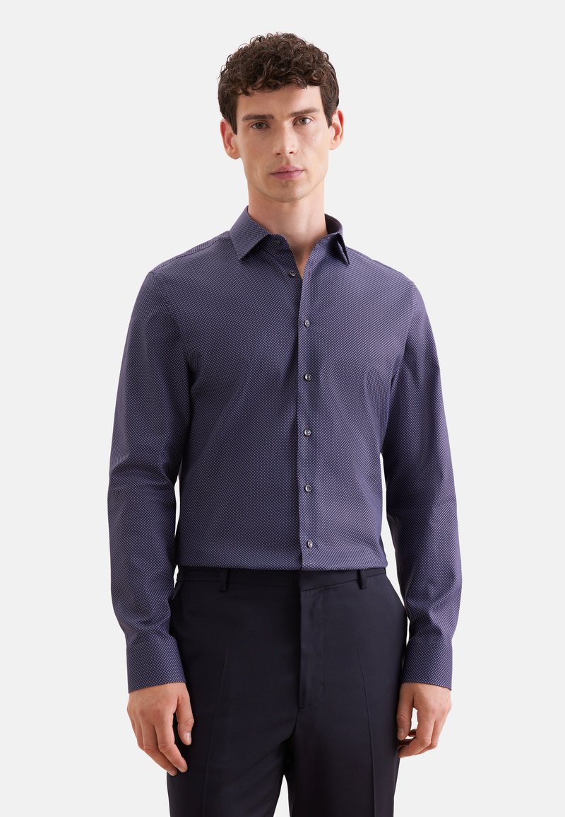 Homme portant une chemise à manches longues boutonnée violet foncé à motifs et un pantalon noir, debout devant un fond clair uni.