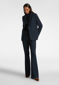 Blazer navy con un solo bottone, revers a freccia e due tasche frontali, abbinato a jeans svasati scuri e scarpe con tacco marroni.