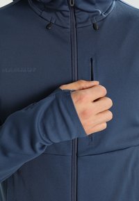 Mørkeblå softshell-jakke med høj krave, lynlåslukning fortil og en indvendig lomme. Har tommelhuller på ærmerne for ekstra dækning.