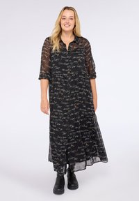 Donna sorridente con lunghi capelli biondi, che indossa un lungo vestito nero con stampa in bianco e stivali neri alla caviglia, in piedi di fronte a uno sfondo bianco.