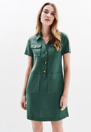 Abito verde a maniche corte con colletto, chiusura frontale a bottoni, due tasche sul petto e due tasche applicate davanti. Realizzato in un tessuto texture.