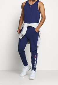 Uomo che indossa una canotta blu navy e pantaloni jogger con scarpe da ginnastica bianche, felpa grigia legata in vita, logo TOMMY SPORT visibile sui pantaloni.