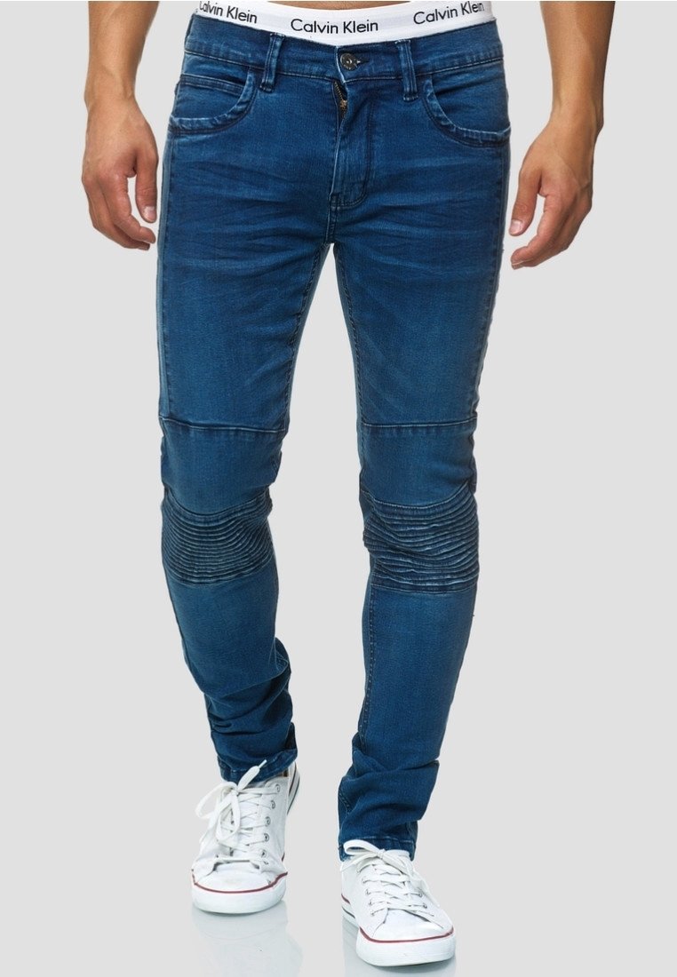 Homme portant un jean slim en denim bleu avec des panneaux côtelés aux genoux, une ceinture blanche Calvin Klein visible et des baskets blanches.