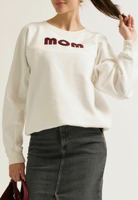 Witte sweatshirt met "Mom" geborduurd in bordeauxrood over de borst, gecombineerd met een donkergrijze spijkerrok. Zachte stof, relaxte pasvorm.