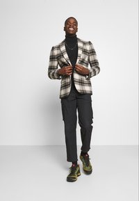 Americana de cuadros en crema, negro y gris, combinada con un jersey de cuello alto negro, pantalones cargo negros y zapatillas de senderismo verdes con detalles rojos.