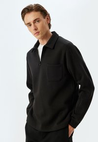 Il pullover nero presenta un collo con zip, una tasca sul petto e una vestibilità rilassata con dettagli delle cuciture a vista. Tessuto dalla trama liscia.