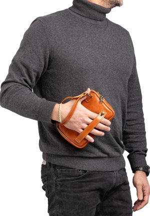 Piccolo borsa rettangolare in pelle color cuoio con un cinturino da polso, chiusura con zip e cuciture a contrasto, tenuta da una persona che indossa un maglione grigio.